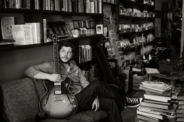 Johnny sitting with his guitar in a room lined with books, black and white