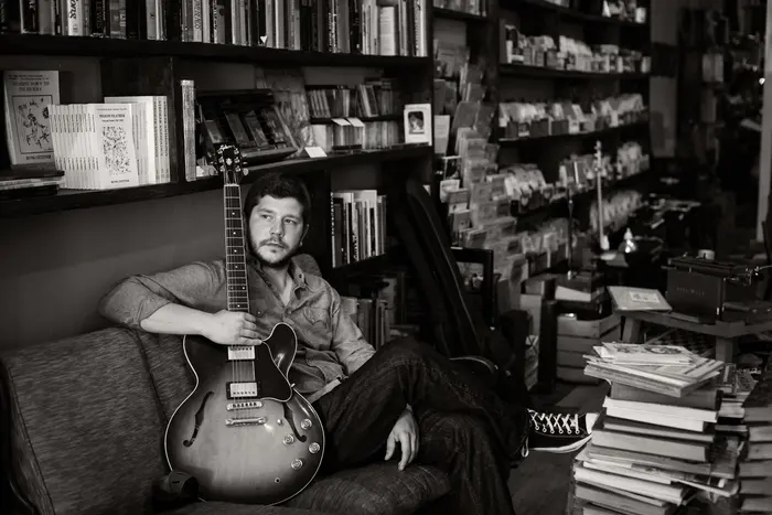 Johnny sitting with his guitar in a room lined with books, black and white