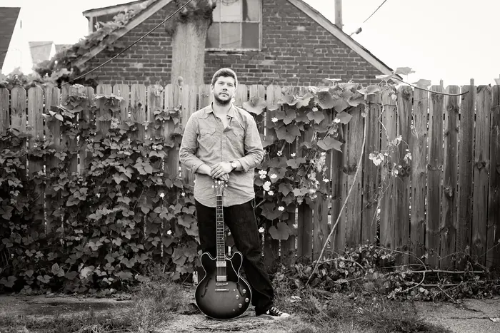 Johnny standing by a wooden fence holding his guitar, black and white