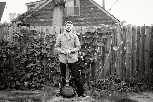Johnny standing by a wooden fence holding his guitar, black and white