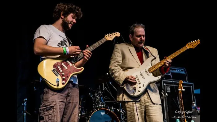 Johnny playing a Stratocaster beside a guitarist in a cream suit
