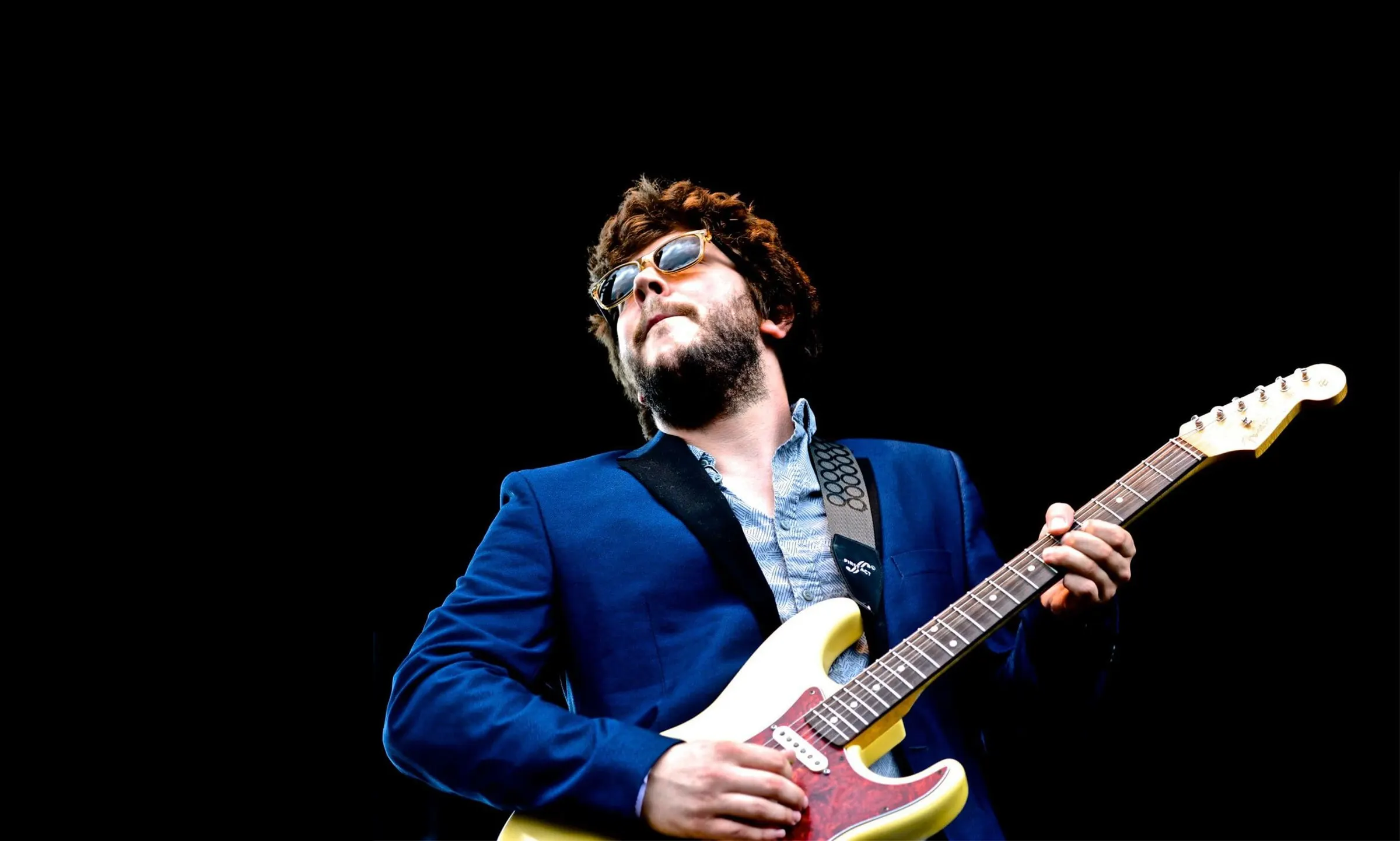 Johnny Rhoades onstage in a blue suit and sunglasses, playing a cream Stratocaster