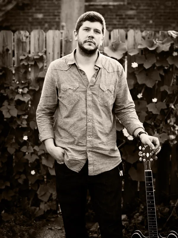 Sepia portrait of Johnny Rhoades standing by a vine-covered fence, guitar at his side