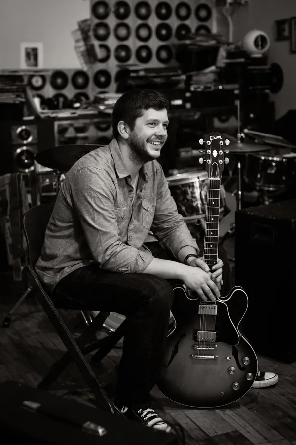 Johnny seated with a hollow-body guitar in front of a wall of amplifiers, black and white